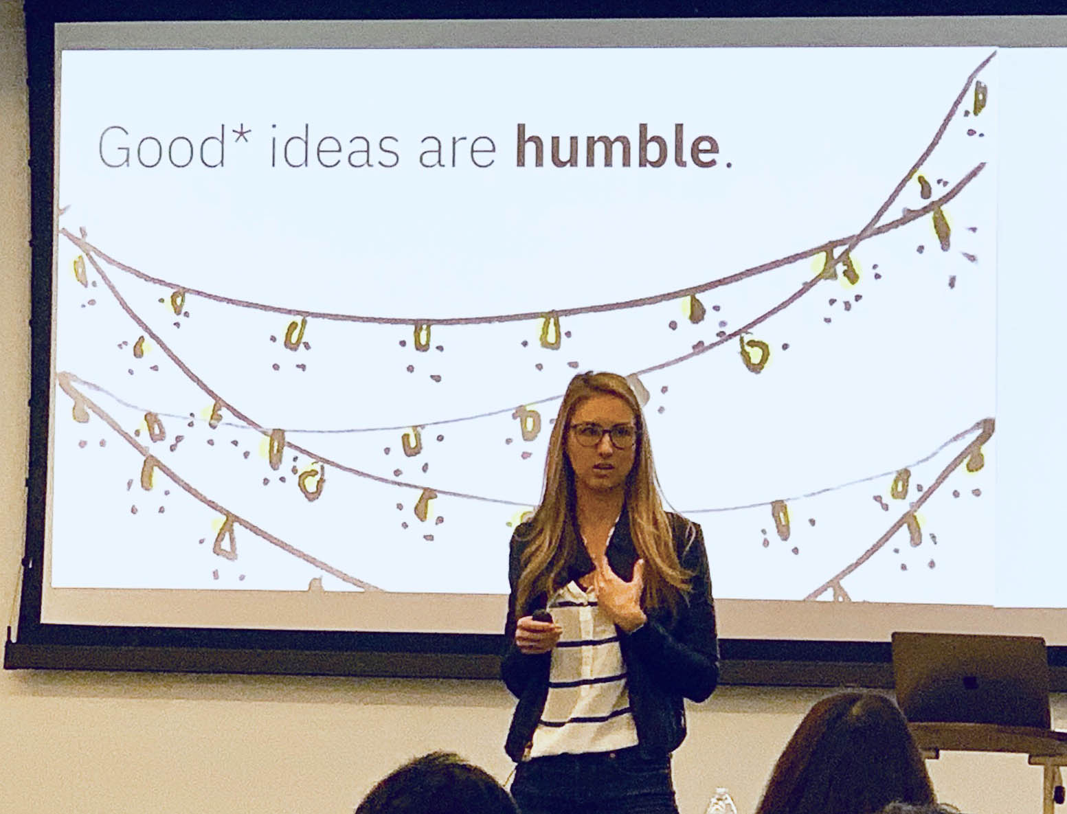 A photo in which I am standing in front of the room at World IA Day, presenting my slide around the concept that “Good* ideas are humble”, which includes an illustrated string of tiny Christmas lights, acting as a metaphor for the humility of a truly good idea: just one amongst many others.